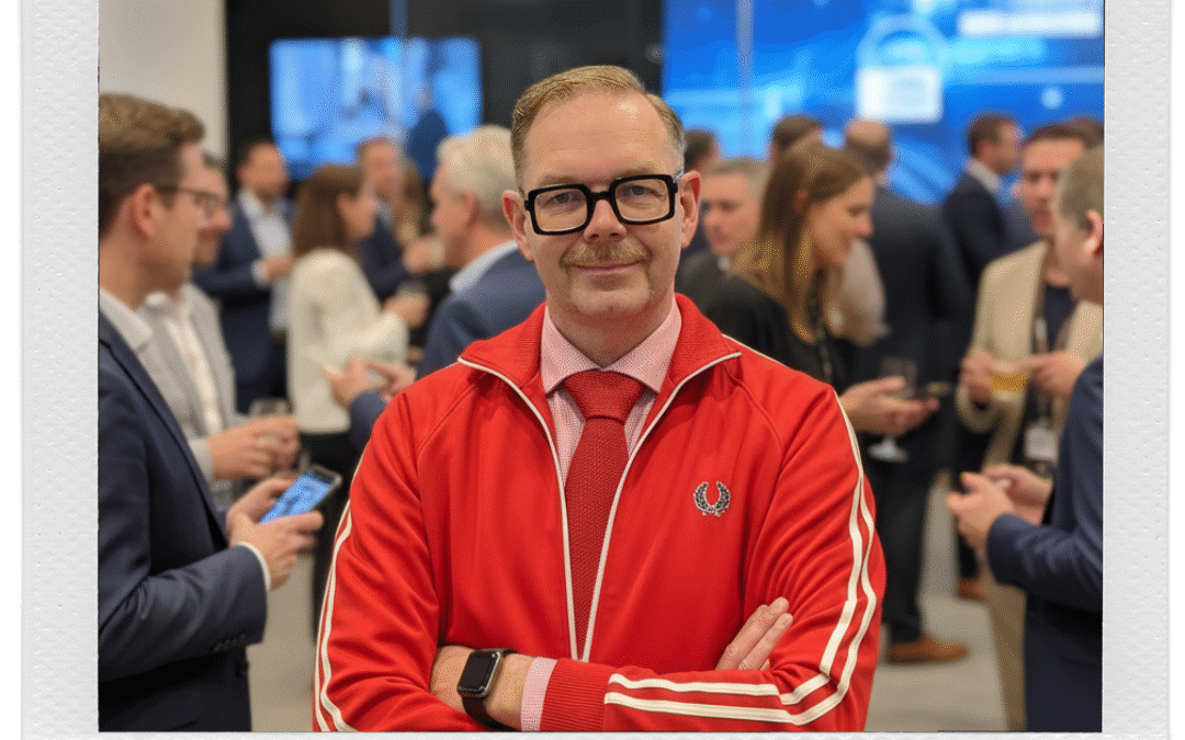 Man med glasögon och mustasch i röd jacka står framför en folksamling på ett nätverksevenemang.