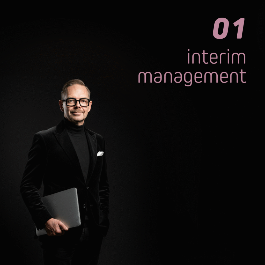 Man i svart kostym med bärbar dator i handen, ler mot kameran. Texten '01 interim management' syns i bakgrunden.