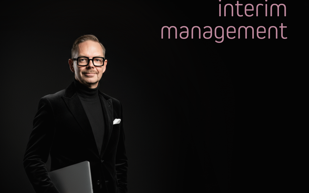 Man i svart kostym med bärbar dator i handen, ler mot kameran. Texten '01 interim management' syns i bakgrunden.