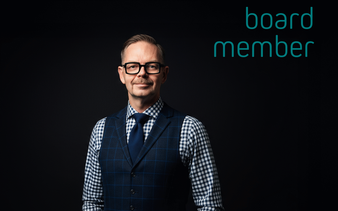 En man i väst och skjorta med glasögon, som står med en laptop i handen och har ett neutralt uttryck.