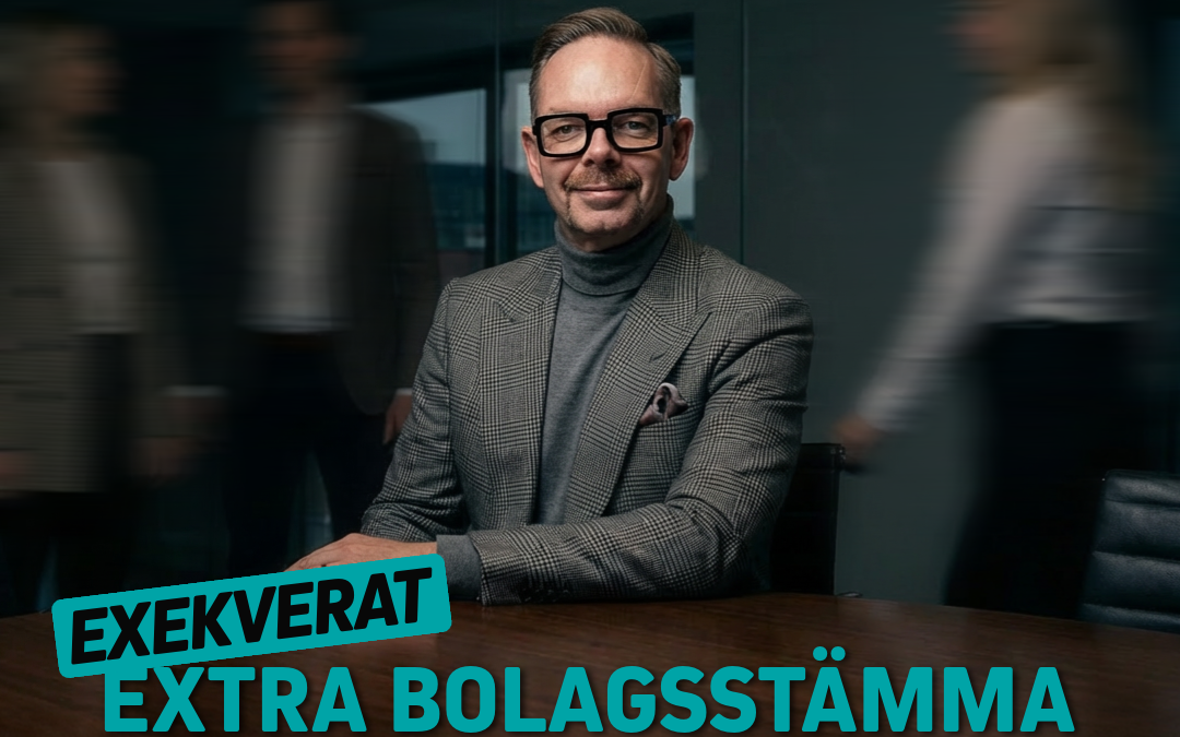 Man i kostym sitter vid ett bord i ett mötesrum med suddiga personer i bakgrunden.